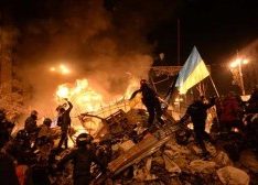 Picture of Ukraine riots in the streets with fire in the background and rioters climbing wreckage with one holding a Ukranian flag