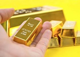 Hand holding 200 gram fine gold bar with stacks of larger gold bars in yellow background