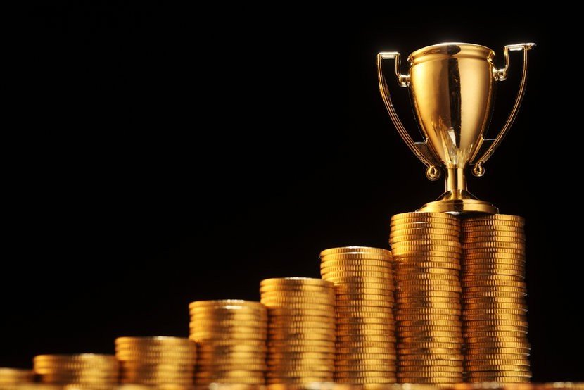 Ascending stacks of gold coins and a gold trophy, against black background