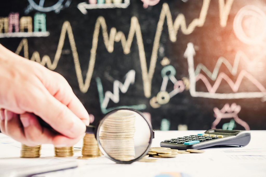 Man hold magnifying glass on stack of gold coins with pile of paperwork as background.