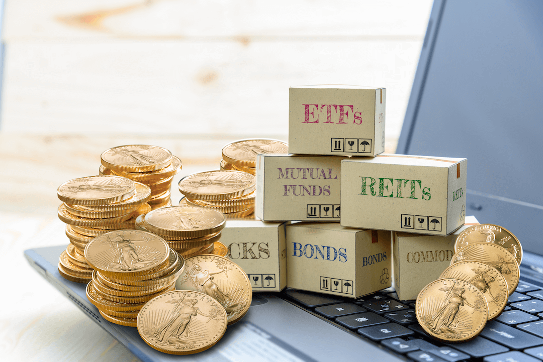 Stacks of gold coins and boxes with stocks, bonds, commodities, REITs, ETFs and mutual funds sitting on top of a laptop keyboard