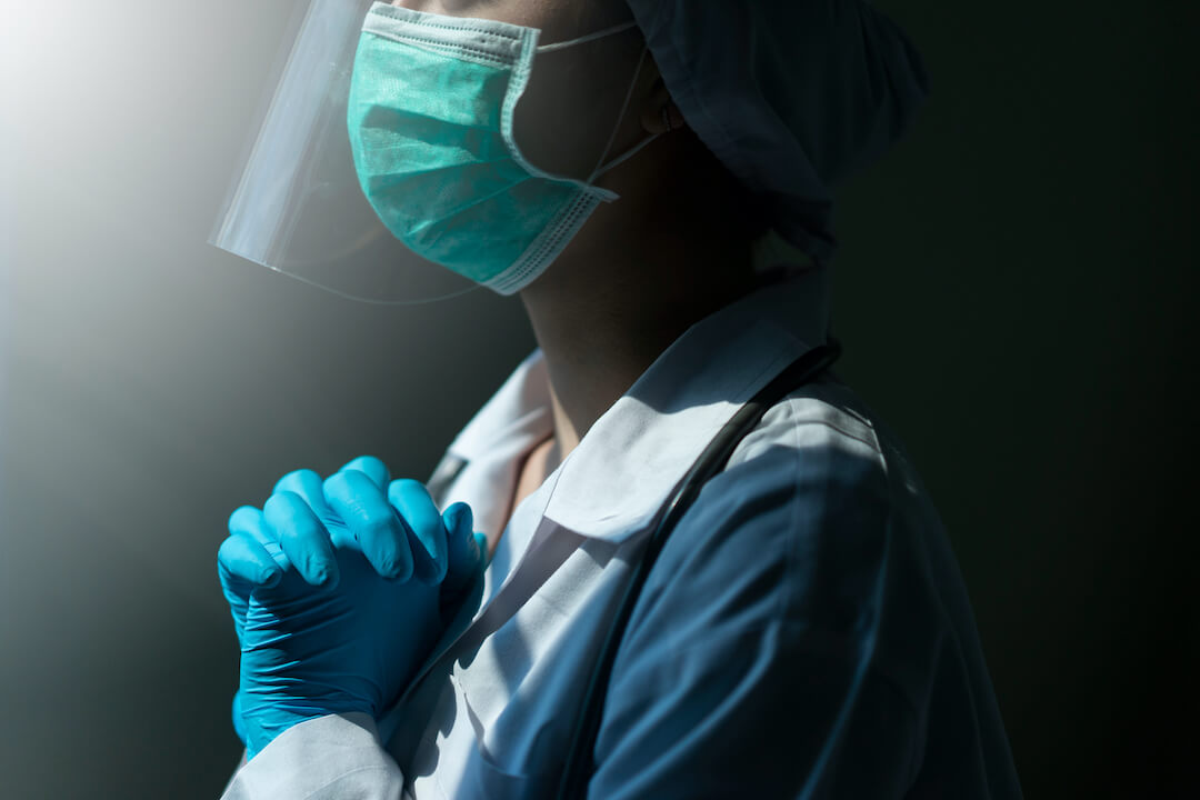 Healthcare worker with mask, shield and surgical gloves on and fingers clasped as if praying