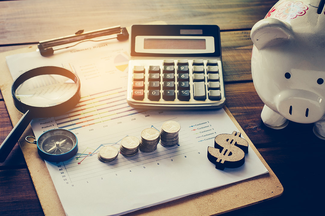 Magnifying glass, compass, stacks of gold coins, a calculator, and dollar symbol sitting on top of a financial summary report, with a piggy bank alongside