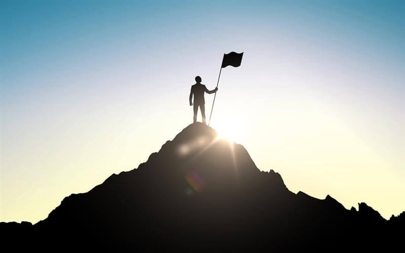 Silhouette of man standing victorious atop a mountain with a flag