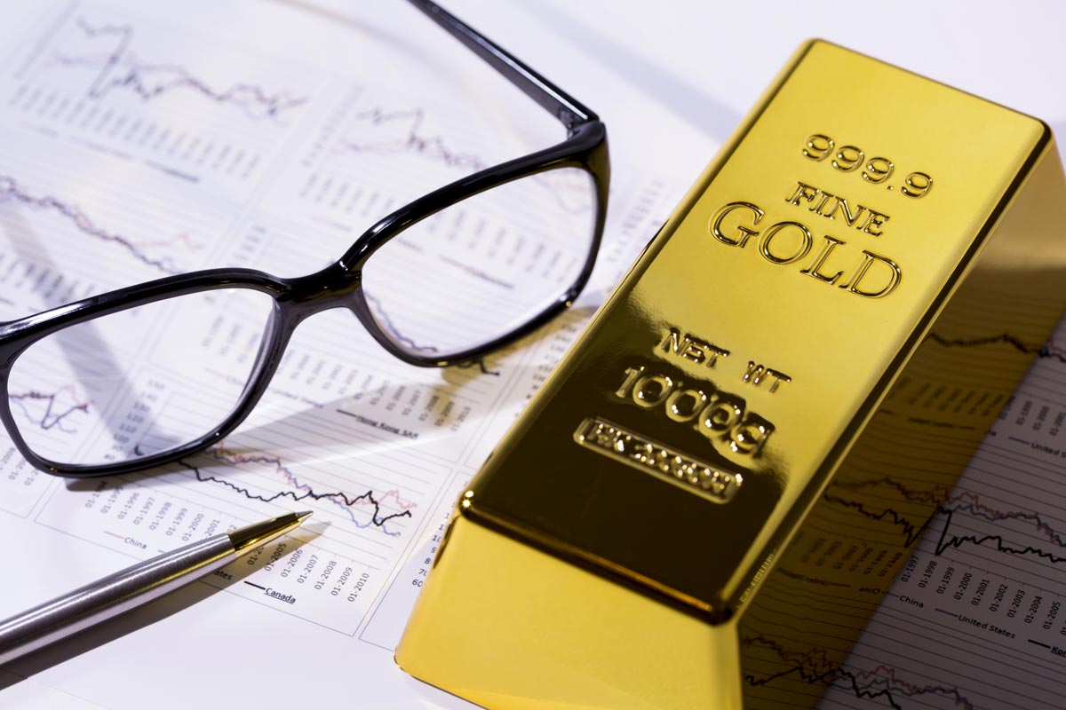 Bar of gold resting on financial charts next to glasses and pen