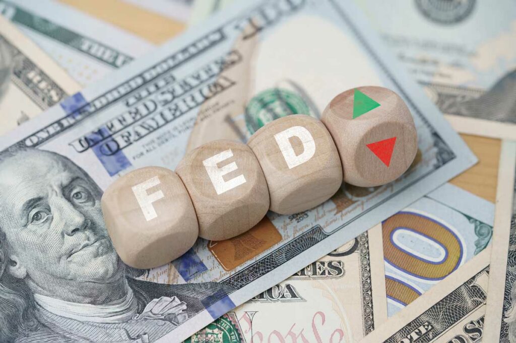Wooden blocks reading "FED" over U.S. currency with fourth block balanced between upward green and downward red arrows
